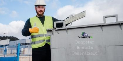 A man in a safety vest and hard hat holding trash next to a commercial dumpster.