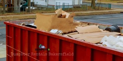 A red residential dumpster containing cardboard boxes and miscellaneous items, situated in a community environment.