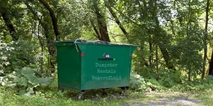 A green yard dumpster placed in the heart of a forest, encircled by dense trees and vibrant greenery.